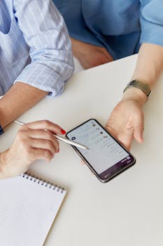 Two adults collaborate using a smartphone and a notepad in an indoor setting.