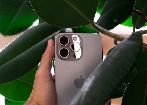 Hand holding a smartphone with triple camera lens against green leaves, showcasing modern design.